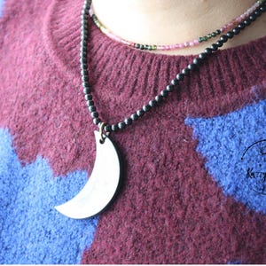 Può includere: Una collana di perline nere con un ciondolo a forma di mezzaluna d'argento. La collana è sovrapposta a una collana di perline rosa e verdi.