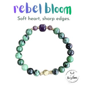 May include: A bracelet featuring green and black speckled beads, purple and gold details, and a central purple square bead. The text "rebel bloom" is above the bracelet, with the words "Soft heart, sharp edges" below.