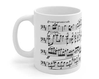 May include: White ceramic coffee mug featuring black musical notes and the text "Quasi improvisando" and "frisolto". The mug has a curved handle and a cylindrical shape. A music-themed gift.