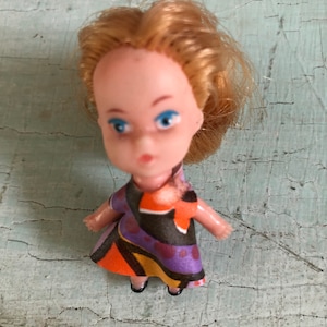 May include: A small plastic doll with blonde hair, blue eyes, and a purple, orange, and black patterned dress.