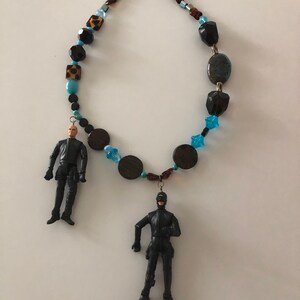 May include: A beaded necklace with two black action figures hanging from it. The necklace features blue, brown, and black beads. One action figure is a scuba diver with flippers. The other action figure is a man in a black suit.