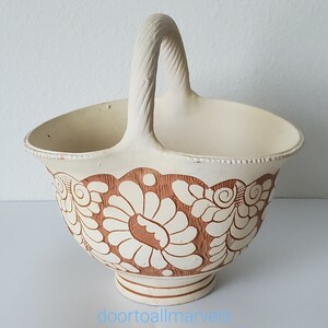 May include: Cream-colored ceramic basket with a woven handle and a floral design in brown. The basket has a scalloped edge and sits on a small pedestal base. The floral pattern is repeated around the basket's exterior.