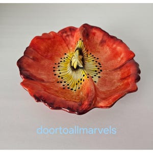 May include: A ceramic dish shaped like a blooming poppy flower with vibrant red petals, a yellow center, and black stamen details. The dish features a textured, organic form with undulating edges and a glossy finish. Text reads doortoallmarvels.