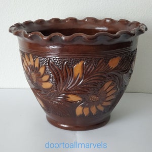 May include: A brown ceramic flower pot with a ruffled rim and carved floral design. The pot features a dark brown glaze with lighter brown accents in the carved details. The text "doortoallmarvels" is at the bottom.