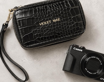 May include: A black rectangular wristlet with a crocodile-embossed texture and a gold-toned zipper. The name VIOLET MAE is printed in gold on the front. Beside it is a black Canon G7X digital camera with a zoom lens and visible text.