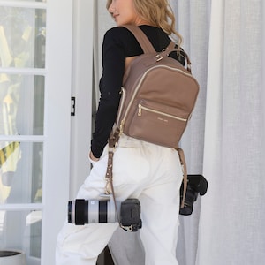 Puede incluir: Una mujer con una camisa negra de manga larga y pantalones blancos está de pie frente a una puerta blanca. Lleva una mochila de cuero marrón con herrajes dorados y una correa de cámara con una cámara adjunta. La cámara es una cámara réflex digital negra con un objetivo grande.