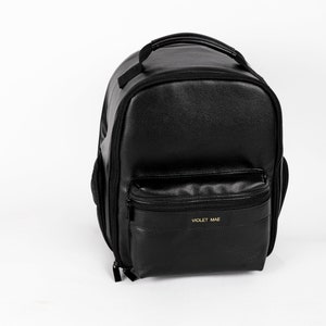 May include: Black leather backpack with a zippered pocket on the front and the text "VIOLET MAE" in gold lettering.