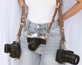 May include: A person wearing a white shirt and blue jeans with two cameras attached to a brown leather camera strap. The camera strap has gold buckles and studs. The cameras are a black digital camera and a silver film camera with the brand name "Minolta" on it.