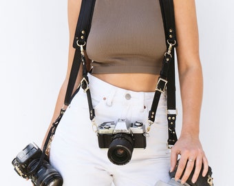 May include: A person wearing a brown tank top and white jeans is using a black leather camera strap to hold two cameras. One camera is a silver Minolta with a black lens, and the other is a black camera with a large white lens.