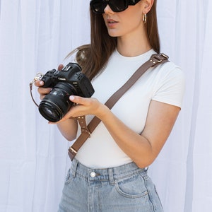 Peut inclure: Une femme portant un t-shirt blanc et un jean bleu tient un appareil photo Canon EOS noir avec une bandoulière en cuir marron. L'appareil photo est équipé d'un objectif.