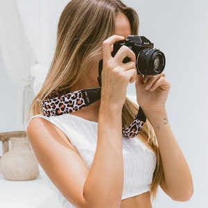 Puede incluir: Una mujer con un top blanco corto y una falda blanca sostiene una cámara negra con una correa de cámara de estampado de leopardo. La correa de la cámara tiene el texto "VOLT TIME" impreso.