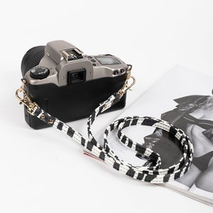 May include: A silver camera with a black and white striped camera strap. The strap is made of leather and has gold hardware.