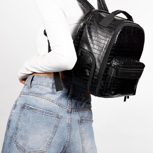 Puede incluir: Mochila negra de piel sint&eacute;tica con textura de piel de cocodrilo. La mochila tiene m&uacute;ltiples compartimentos con cremallera, un asa superior y correas ajustables para los hombros. La bolsa se lleva sobre los hombros. La bolsa tiene forma rectangular.