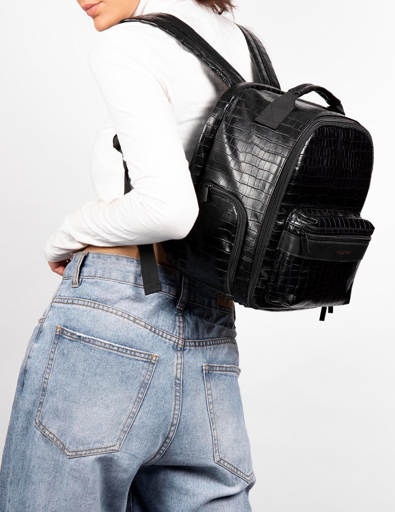 Puede incluir: Mochila negra de piel sint&eacute;tica con textura de piel de cocodrilo. La mochila tiene m&uacute;ltiples compartimentos con cremallera, un asa superior y correas ajustables para los hombros. La bolsa se lleva sobre los hombros. La bolsa tiene forma rectangular.