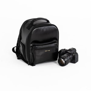 May include: Black leather camera backpack with a gold logo that says "Violet Mae". A black camera with a lens is in front of the backpack.