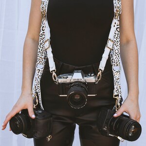 May include: A white and brown leopard print camera strap with gold hardware. The strap is worn by a person holding three cameras, including a silver Minolta X-3000 camera.