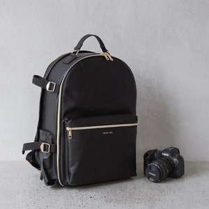 May include: Black backpack with gold-colored zippers and trim, and a top handle. The backpack has a front pocket with the text "VIOLET MAE". A black camera with a lens is next to the backpack.