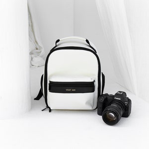 May include: White camera backpack with black trim and the text "VIOLET MAE" on the front. A black camera with a lens is in front of the backpack.