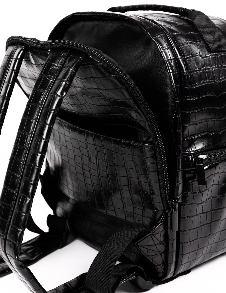 Puede incluir: Mochila negra con textura de piel de cocodrilo. La bolsa tiene un asa superior, correas ajustables y m&uacute;ltiples compartimentos con cremallera. El interior es negro y la bolsa est&aacute; abierta.