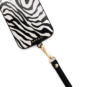 Peut inclure: Coque de smartphone avec un motif zébré noir et blanc. L'étui a un bord noir et un fermoir doré attaché à une lanière noire. La marque "BURGA" est imprimée sur l'étui.