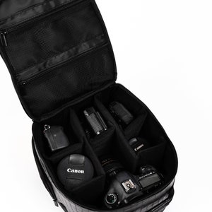 Puede incluir: Bolsa de c&aacute;mara negra con m&uacute;ltiples compartimentos, abierta para revelar varios equipos de c&aacute;mara, incluyendo una c&aacute;mara Canon, lente y accesorios. La bolsa tiene un exterior texturizado y bolsillos con cremallera.