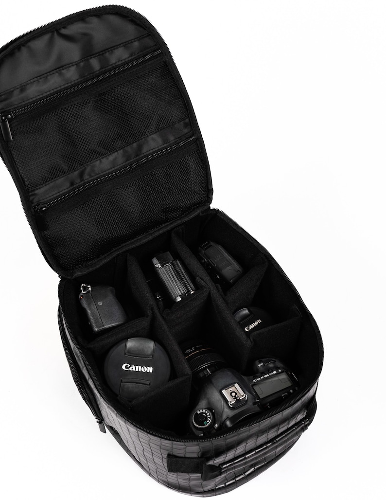 Puede incluir: Bolsa de c&aacute;mara negra con m&uacute;ltiples compartimentos, abierta para revelar varios equipos de c&aacute;mara, incluyendo una c&aacute;mara Canon, lente y accesorios. La bolsa tiene un exterior texturizado y bolsillos con cremallera.