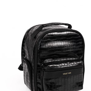 Puede incluir: Mochila negra con textura de piel de cocodrilo. Cuenta con asa superior, bolsillo frontal y bolsillo lateral. El bolsillo frontal tiene el texto "VIOLET MAE" en letras doradas. La mochila tiene cierre de cremallera y correas ajustables.
