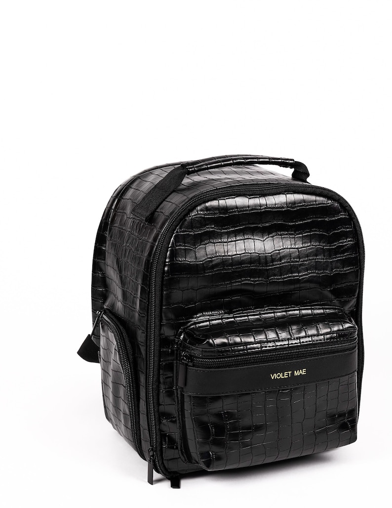 Puede incluir: Mochila negra con textura de piel de cocodrilo. Cuenta con asa superior, bolsillo frontal y bolsillo lateral. El bolsillo frontal tiene el texto "VIOLET MAE" en letras doradas. La mochila tiene cierre de cremallera y correas ajustables.