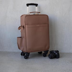 May include: A brown rolling suitcase with a retractable handle and four wheels. A black camera is next to the suitcase. The suitcase has a side pocket and a small bow detail. The suitcase is on a grey surface.