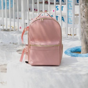 Puede incluir: Una mochila rosa claro con cremalleras y detalles dorados. La mochila tiene un bolsillo frontal y correas ajustables. La bolsa está sobre una superficie blanca, con una valla blanca y un árbol al fondo.