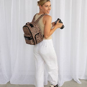 Puede incluir: Una mujer con pantalones blancos y zapatillas lleva una mochila de estampado de leopardo y una cámara negra.
