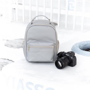 Puede incluir: Una mochila gris para cámara con detalles dorados y una cámara negra con lente sobre una superficie blanca con la palabra "ASSOU" escrita en ella.