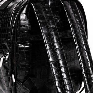Puede incluir: Mochila negra con textura de piel de cocodrilo. Cuenta con asa superior, correas ajustables y cierre de cremallera. Fabricada en material de polipiel brillante.