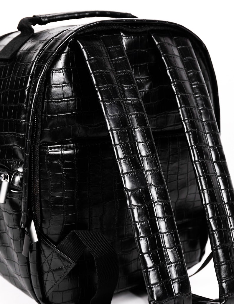Puede incluir: Mochila negra con textura de piel de cocodrilo. Cuenta con asa superior, correas ajustables y cierre de cremallera. Fabricada en material de polipiel brillante.