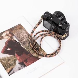 May include: A black camera with a brown and black patterned strap. The camera is lying on a magazine with a photo of a woman wearing a red suit.