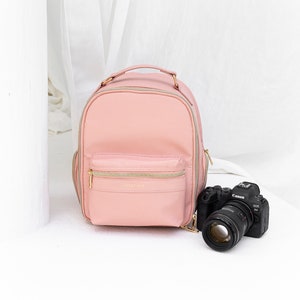 Könnte beinhalten: Ein rosa Leder-Kamerarucksack mit einem goldenen Reißverschluss und dem Text "Voyage & Me" auf der Vorderseite. Eine schwarze Canon EOS 6D Kamera mit einem Objektiv befindet sich vor dem Rucksack.