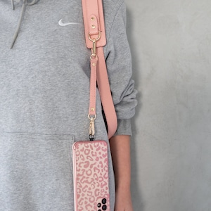 May include: A pink leather phone strap with gold-colored hardware is attached to a phone case with a pink leopard print design. The strap is worn over the shoulder. The phone case is a light pink color.
