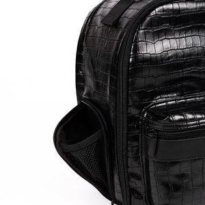 Puede incluir: Mochila negra con textura brillante de piel de cocodrilo. Cuenta con un asa superior, un compartimento principal con cremallera y un bolsillo frontal. Incluye un bolsillo lateral con interior de malla. La bolsa tiene un dise&ntilde;o elegante y moderno.