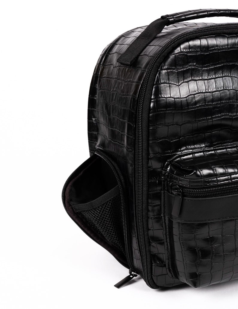 Puede incluir: Mochila negra con textura brillante de piel de cocodrilo. Cuenta con un asa superior, un compartimento principal con cremallera y un bolsillo frontal. Incluye un bolsillo lateral con interior de malla. La bolsa tiene un dise&ntilde;o elegante y moderno.