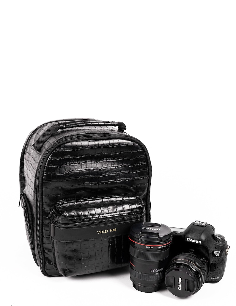 Puede incluir: Mochila negra para c&aacute;mara con textura de piel de cocodrilo, una c&aacute;mara Canon EOS 5D Mark III y dos objetivos. La mochila tiene el texto "VIOLET MAE" en la parte delantera. Las lentes tienen detalles en negro y rojo.