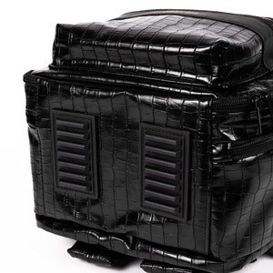 Puede incluir: Mochila negra con textura brillante de piel de cocodrilo. La bolsa presenta dos parches rectangulares texturizados y un compartimento con cremallera. El dise&ntilde;o es elegante y moderno, con un enfoque en el material texturizado y la forma.