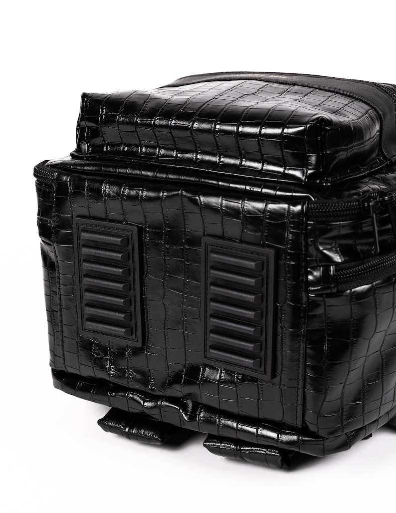 Puede incluir: Mochila negra con textura brillante de piel de cocodrilo. La bolsa presenta dos parches rectangulares texturizados y un compartimento con cremallera. El dise&ntilde;o es elegante y moderno, con un enfoque en el material texturizado y la forma.