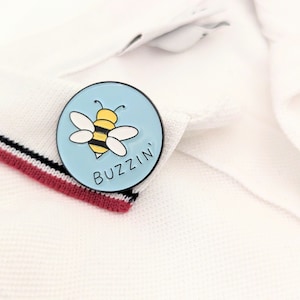 May include: A blue and white enamel pin with a cartoon bee and the text "Buzzin'" on a white polo shirt.