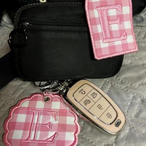 May include: A black nylon belt bag featuring two pink and white gingham embroidered patches, each displaying the letter E. One patch is rectangular with rounded corners, and the other is circular with a scalloped edge. A beige car key fob is attached.