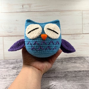 May include: A crocheted blue owl with purple wings and white eyes. The owl has an orange beak and is closed-eyed.