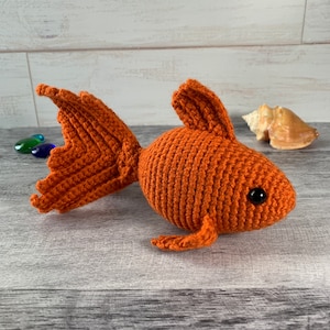 May include: A crocheted orange goldfish toy with black eyes. The toy is sitting on a light wood surface.
