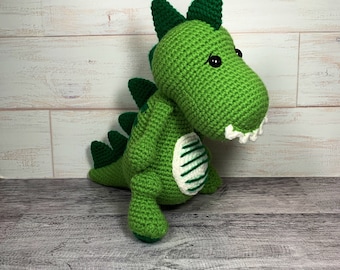 May include: Green crocheted dinosaur toy with white stripes on its back. The dinosaur has black eyes and a toothy grin.