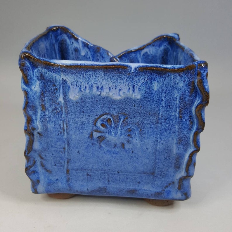 Pu&ograve; includere: Un vaso quadrato in ceramica fatto a mano con una vivace glassa blu e un bordo strutturato e ondulato. Il vaso presenta un motivo decorativo in rilievo sul davanti e poggia su piccoli piedini arrotondati. La glassa ha un effetto bianco gocciolato.
