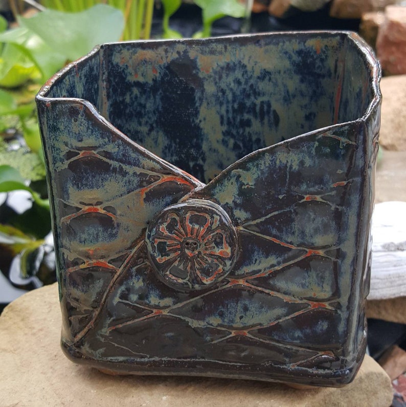 Pu&ograve; includere: Un vaso quadrato in ceramica con smalto blu scuro e verde. Il vaso presenta un motivo strutturato e un bottone decorativo con un motivo floreale. Il vaso &egrave; alto circa 15 cm e largo 13 cm.