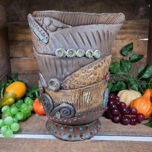 May include: A handcrafted, brown ceramic vase with a textured surface and decorative embellishments. The vase features layers with turquoise, red, and green accents. The vase is approximately 12 inches tall and has a unique, artistic design.